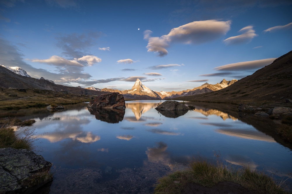 Photo d'un paysage de montagne.
Le Cervin se reflète dans un lac. Deux gros rochers encadrent le reflet de la montagne. Le ciel est bleu mais de nombreux nuages lenticulaires, ressemblant à des soucoupes volantes, l'ont envahi. Ces nuages se reflètent aussi dans le lac. Droit au-dessus de la montagne, on distingue la pleine lune, toute petite.
Le lac est plongé dans l'ombre, les montagnes les plus hautes sont déjà éclairées par le soleil.
-
Photo of a mountain landscape.
The Matterhorn is reflected in a lake. Two large rocks frame the mountain's reflection. The sky is blue, but numerous lenticular clouds, resembling flying saucers, have invaded it. These clouds are also reflected in the lake. High above the mountain, we can make out the tiny full moon.
The lake is plunged into shadow, the higher mountains already lit up by the sun.