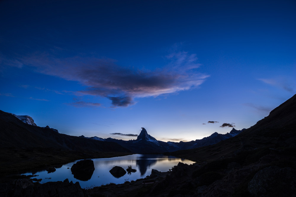 Photo d'un paysage de montagne au crépuscule.
Le Cervin se reflète dans un petit lac de montagne d'où émergent quelques rochers. Il fait sombre, le lac fait une tache de lumière dans le paysage.
Le ciel occupe les deux tiers supérieurs de l'image, il est bleu, avec un petite lueur jaune près de l'horizon. Un nuage vaguement rose occupe le centre du ciel, juste au-dessus du Cervin.
-
Photo of a mountain landscape at dusk.
The Matterhorn is reflected in a small mountain lake from which a few rocks emerge. It's dark, and the lake forms a spot of light in the landscape.
The sky occupies the upper two-thirds of the image; it is blue, with a small yellow glow near the horizon. A vaguely pink cloud occupies the center of the sky, just above the Matterhorn.