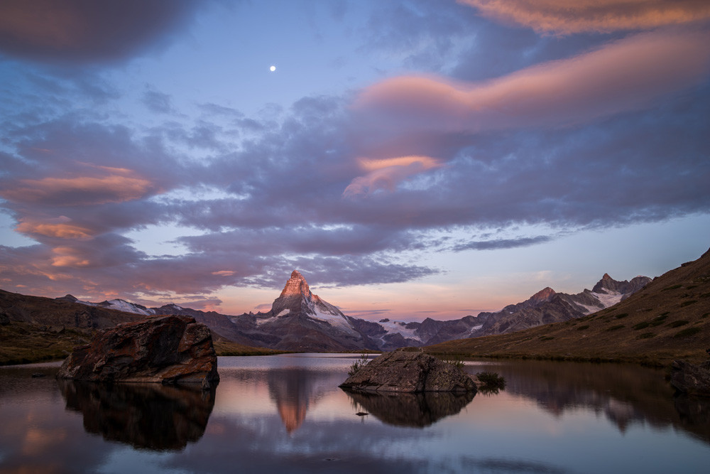 Photo d'un paysage de montagne au lever du soleil. L'image est au format paysage.
Le Cervin se reflète dans un petit lac de montagne, d'où émergent deux gros rochers. Le reflet est légèrement trouble.
Le sommet du Cervin est le seul éclairé par le soleil, presque rouge. Le ciel juste derrière est rose.
Le reste du ciel est encombré de nuages aplatis, virant du gris au rose. Droit au-dessus du Cervin, on devine la pleine lune.
-
Photo of a mountain landscape at sunrise. The image is in landscape format.
The Matterhorn is reflected in a small mountain lake, from which two large rocks emerge. The reflection is slightly cloudy.
The summit of the Matterhorn is the only one lit by the sun, almost red. The sky just behind is pink.
The rest of the sky is cluttered with flattened clouds, turning from grey to pink. Directly above the Matterhorn, we can make out the full moon.