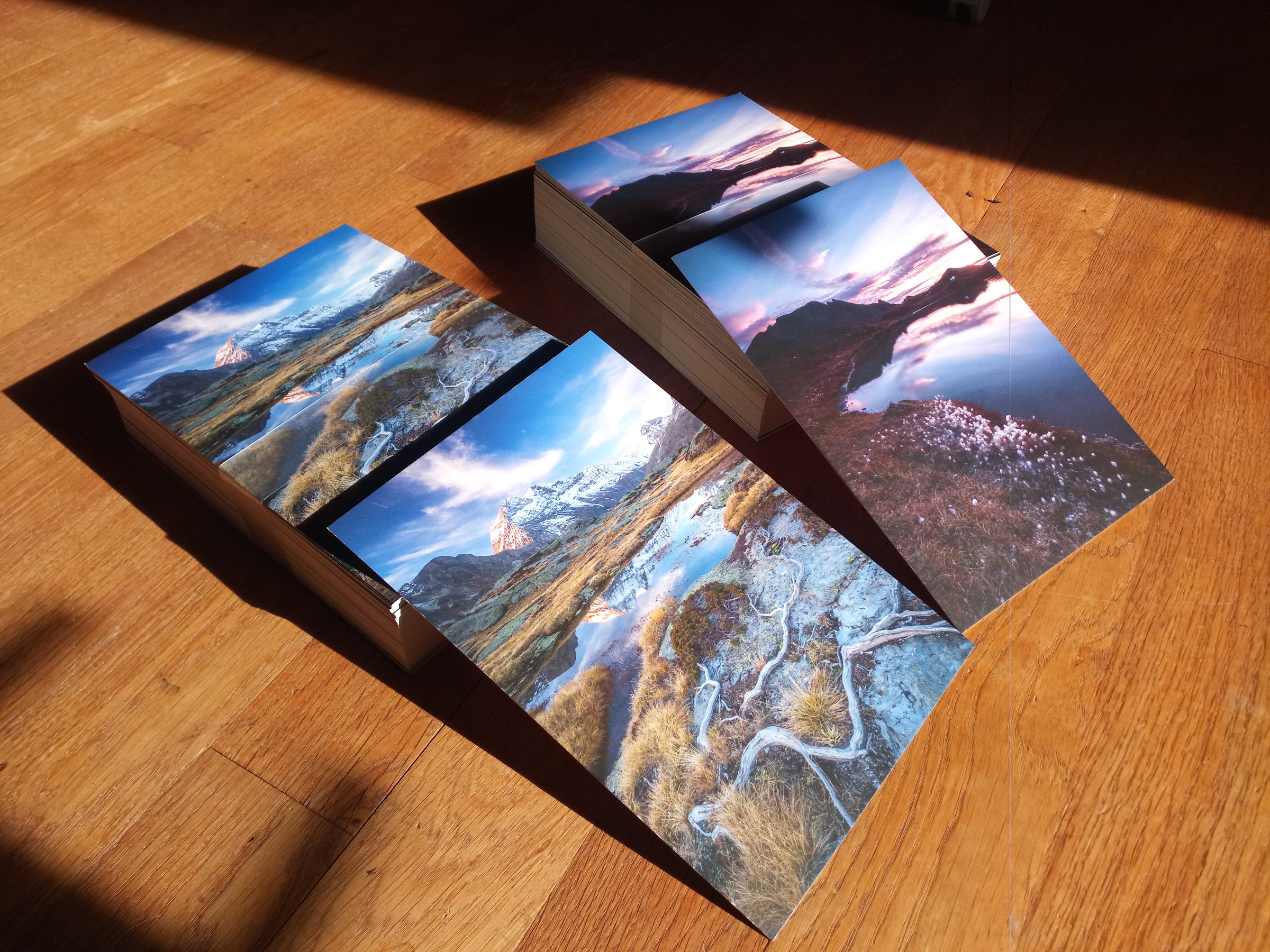 Photo de deux piles de cartes postales posées sur un parquet en bois. 
Les cartes montrent chacune un paysage de montagne différent au format vertical. Celle de gauche montre une montagne enneigée dominant un paysage de lande avec un petit lac et des racines au premier plan, celle de droite la berge d'un petit lac de montagne, couverte de linaigrettes, au lever du soleil.