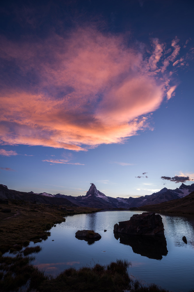 Photo d'un paysage de montagne au coucher du soleil.
Un gros nuage rose domine un petit lac de montagne plongé dans l'ombre. Il y a deux gros rochers dans le lac, et à l'horizon on reconnait la forme élancée du Cervin.
-
Photo of a mountain landscape at sunset.
A large pink cloud dominates a small mountain lake plunged into shadow. There are two large rocks in the lake, and on the horizon you can see the slender shape of the Matterhorn.