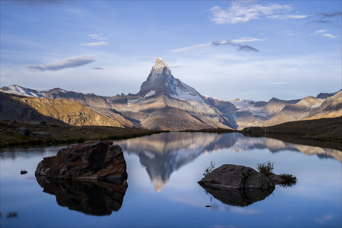 Photo d'un paysage de montagne.
Le Cervin se reflète dans un lac, le reflet est relativement net.
Dans le lac, deux gros rochers émergent de l'eau de part et d'autre de la montagne.
Le lac est dans l'ombre, mais les montagnes sont éclairées par intermittence, du fait de la présence de nuages dans le ciel.
-
Photo of a mountain landscape.
The Matterhorn is reflected in a lake; the reflection is relatively clear.
In the lake, two large rocks emerge from the water on either side of the mountain.
The lake is in shadow, but the mountains are intermittently illuminated by the clouds in the sky.