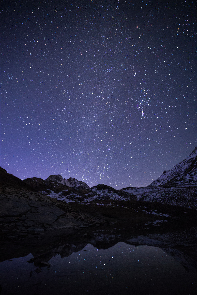 Photo d'un paysage de montagne sous les étoiles, au format portrait.
Un ciel très étoilé se reflète dans un lac bordé de montagnes. Les pentes sont saupoudrées de neige, quelques étoiles se reflètent dans le lac.
Le ciel occupe les deux tiers supérieurs de l'image.
-
Portrait photo of a mountain landscape under the stars.
A starry sky is reflected in a lake surrounded by mountains. The slopes are dusted with snow, and a few stars are reflected in the lake.
The sky occupies the upper two-thirds of the image.