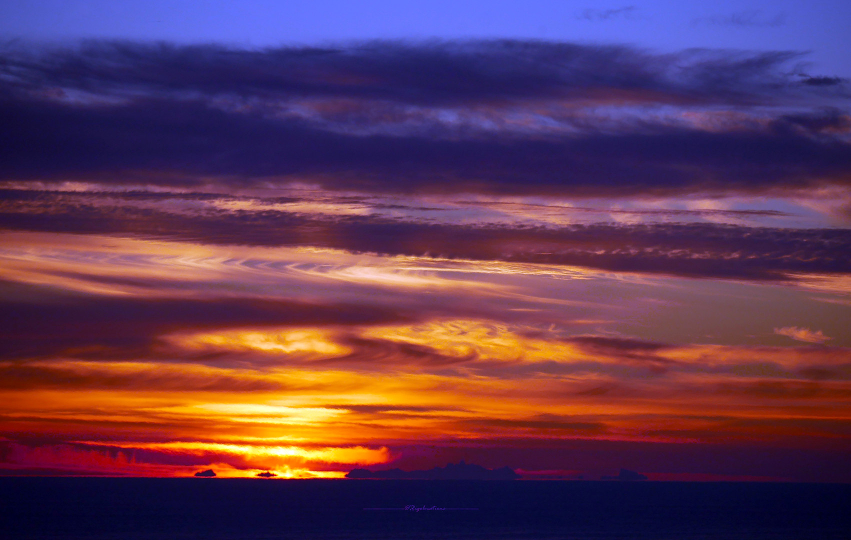 Photo du soleil en train de commencer à apparaitre derrière la ligne d'horizon, sur la mer. Le long de la ligne d'horizon, on aperçoit ce qui ressemble à une île. Il s'agit en réalité d'un phénomène de réfraction lumineuse qui fait que l'on peut voir, comme pour un mirage, le sommet des montagnes Corses depuis le rivage du Continent. Au dessus de l'horizon, des nuages sous forme de bandes cotonneuses sont illuminés d'or et d'orange là où le soleil va apparaitre. Au dessus, le reste des nuages se colore de rose, doré, mauve et violet foncé. On aperçoit un peu de ciel bleu-mauve au dessus de la bande nuageuse.