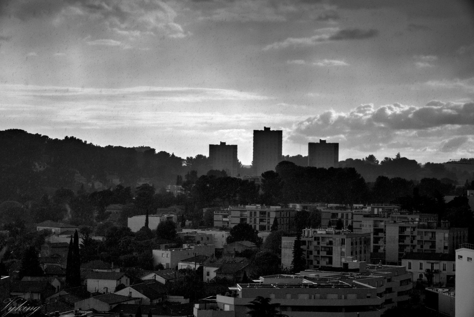 Photographie en noir et blanc très contrastée prise du centre vers l'ouest de Nîmes, sous la pluie. La durée d’exposition étant très courte, chaque goûte de pluie fait une tache la faisant ressembler à un flocon, ou à une pluie de cendre, effet renforcé par les jeux d’ombre et de lumière créés par le soleil et les nuages, ainsi que par les trois tours HLM semblant sortir d'une sorte de brouillard en arrière plan, donnant une dimension apocalyptique à la scène.