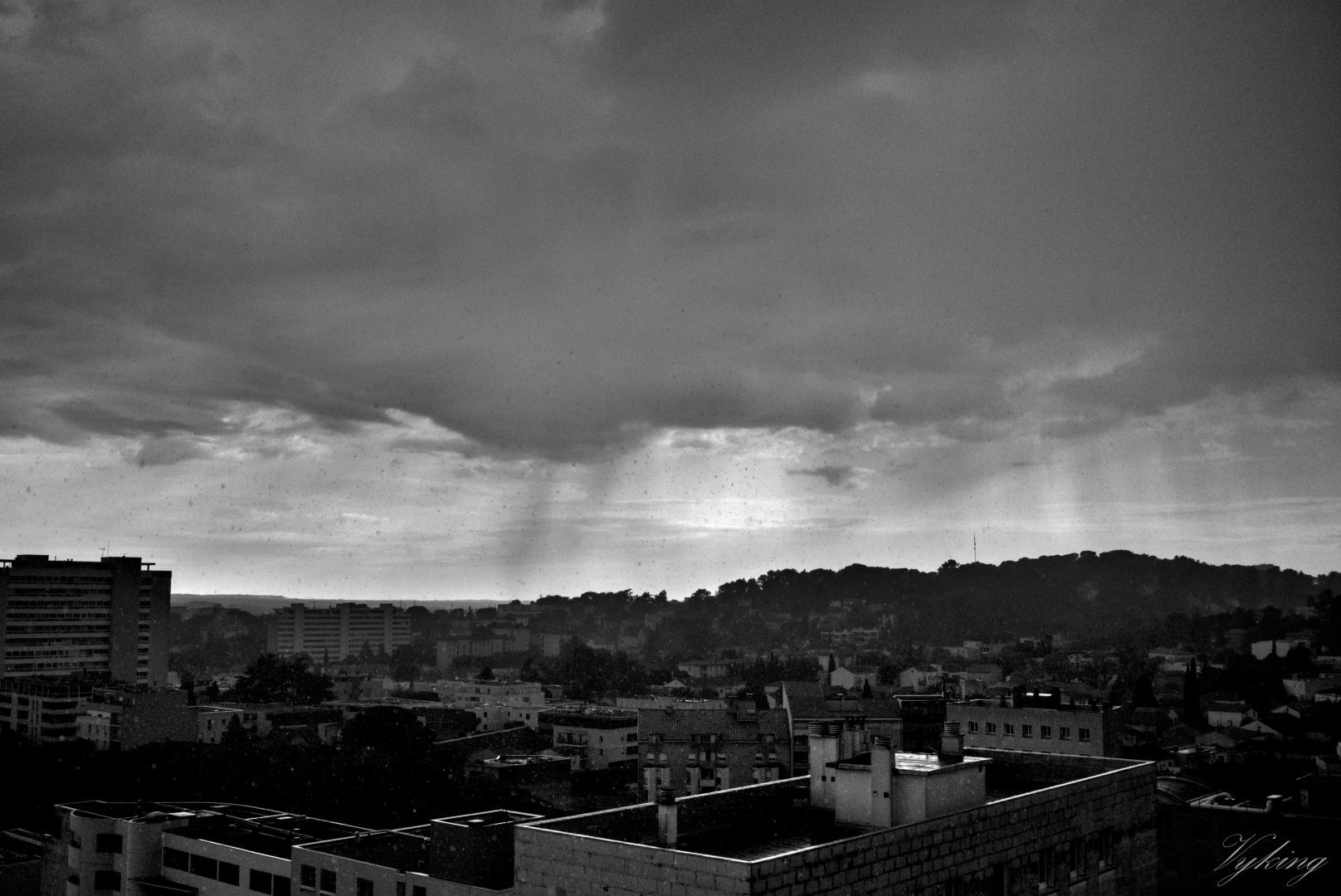 Photographie en noir et blanc très contrastée prise du centre vers le sud-ouest de Nîmes, sous la pluie. La durée d’exposition étant très courte, chaque goûte de pluie fait une tache la faisant ressembler à un flocon, ou à une pluie de cendre, effet renforcé par les jeux d’ombre et de lumière créés par le soleil et les nuages, donnant une dimension apocalyptique à la scène.