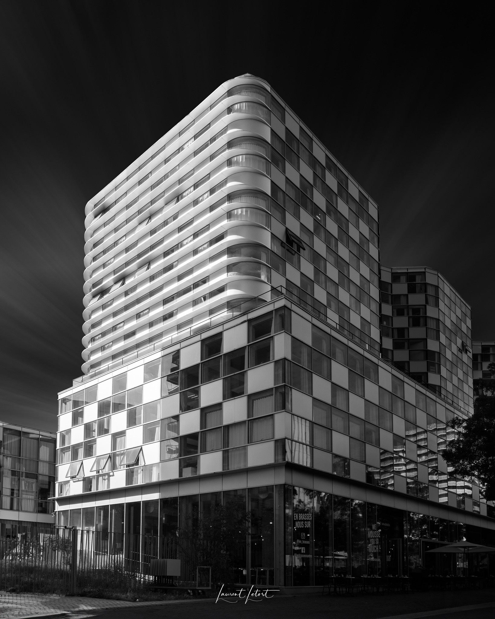 🇫🇷 Un bâtiment moderne de plusieurs étages avec des balcons incurvés et du verre réfléchissant, capturé dans un noir et blanc saisissant.
🇬🇧 A modern, multi-story building with curved balconies and reflective glass, captured in striking black and white.