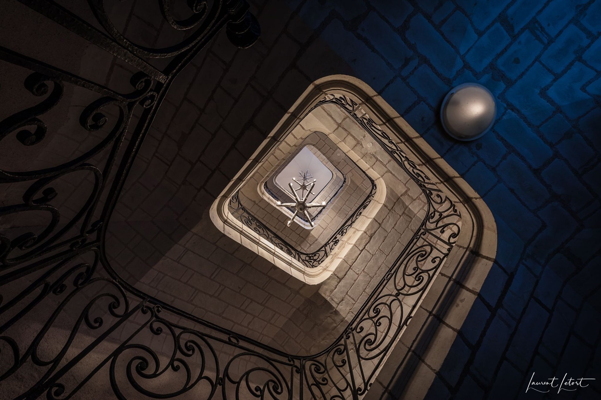 🇫🇷 Vue descendante d'un escalier en colimaçon doté de rampes en fer ornées et d'un lustre moderne sur un mur texturé et doucement éclairé.

🇬🇧 A downward view of a spiral staircase featuring ornate iron railings and a modern chandelier against a softly lit, textured wall.