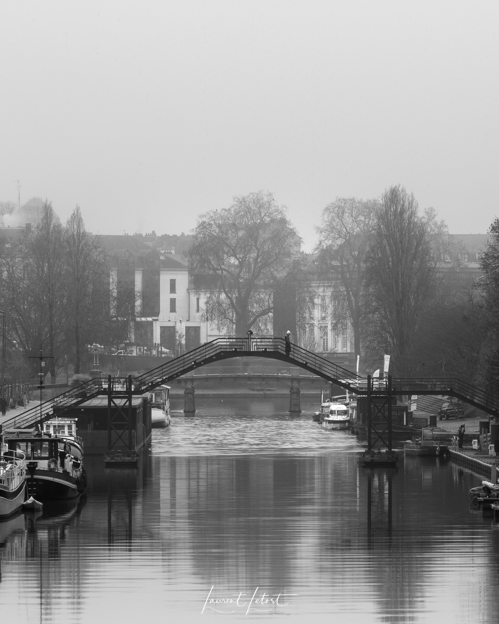 🇫🇷 Scène de rivière brumeuse avec une passerelle reliant le quai Barbusse à l'ile Versailles à Nantes, des bateaux alignés le long de la rive et des arbres dénudés, créant une atmosphère sereine et monochrome.

🇬🇧  Misty river scene with a footbridge linking quai Barbusse to Ile Versailles in Nantes, boats lined up along the bank and bare trees, creating a serene, monochrome atmosphere.