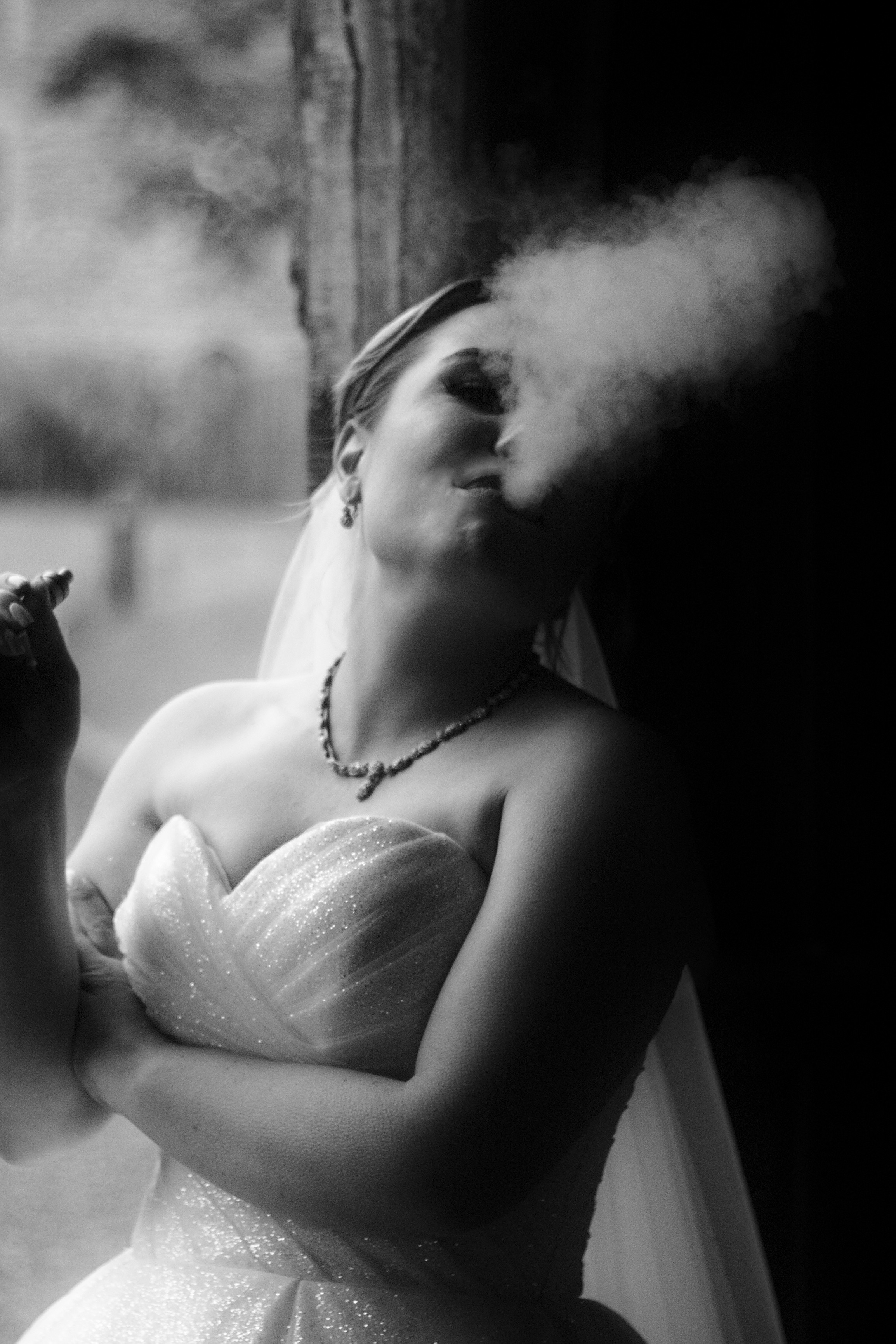 Photo en noir et blanc d'une femme blanche en tenue de mariée : une robe bustier blanche scintillante, un voile dans les cheveux, et un collier de gemmes assorti à ses boucles d'oreille. Elle croise les bras, adossée à l'entrée d'une grange en bois, et tourne la tête pour souffler vers le haut la fumée de sa vapoteuse.