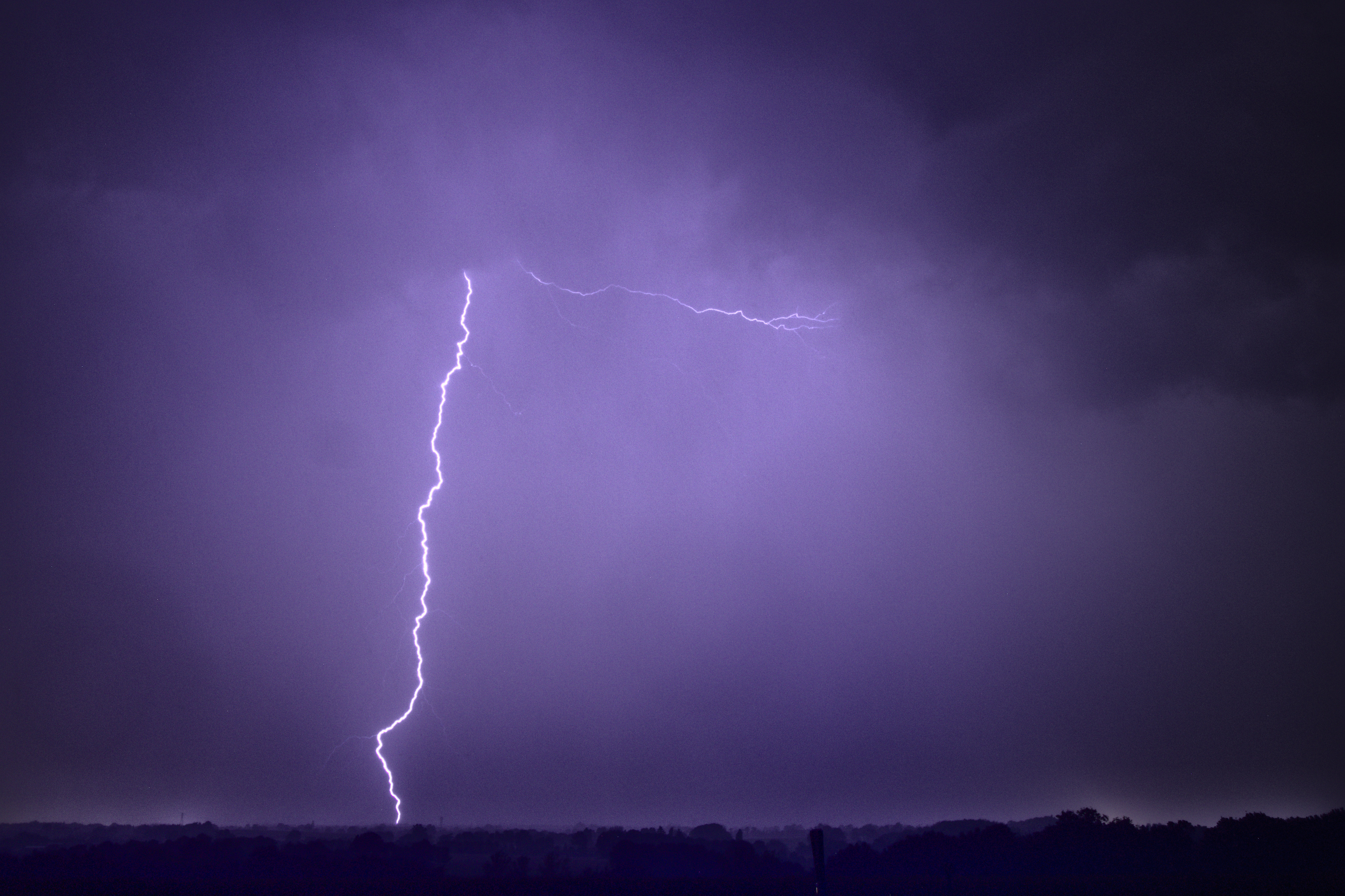 Photo de nuit d'un impact de foudre tout droit, frappant un point à l'horizon derrière un paysage de bocage. L'éclair illumine l'atmosphère de violet, et une unique ramification part de la base du nuage, presque en angle droit.