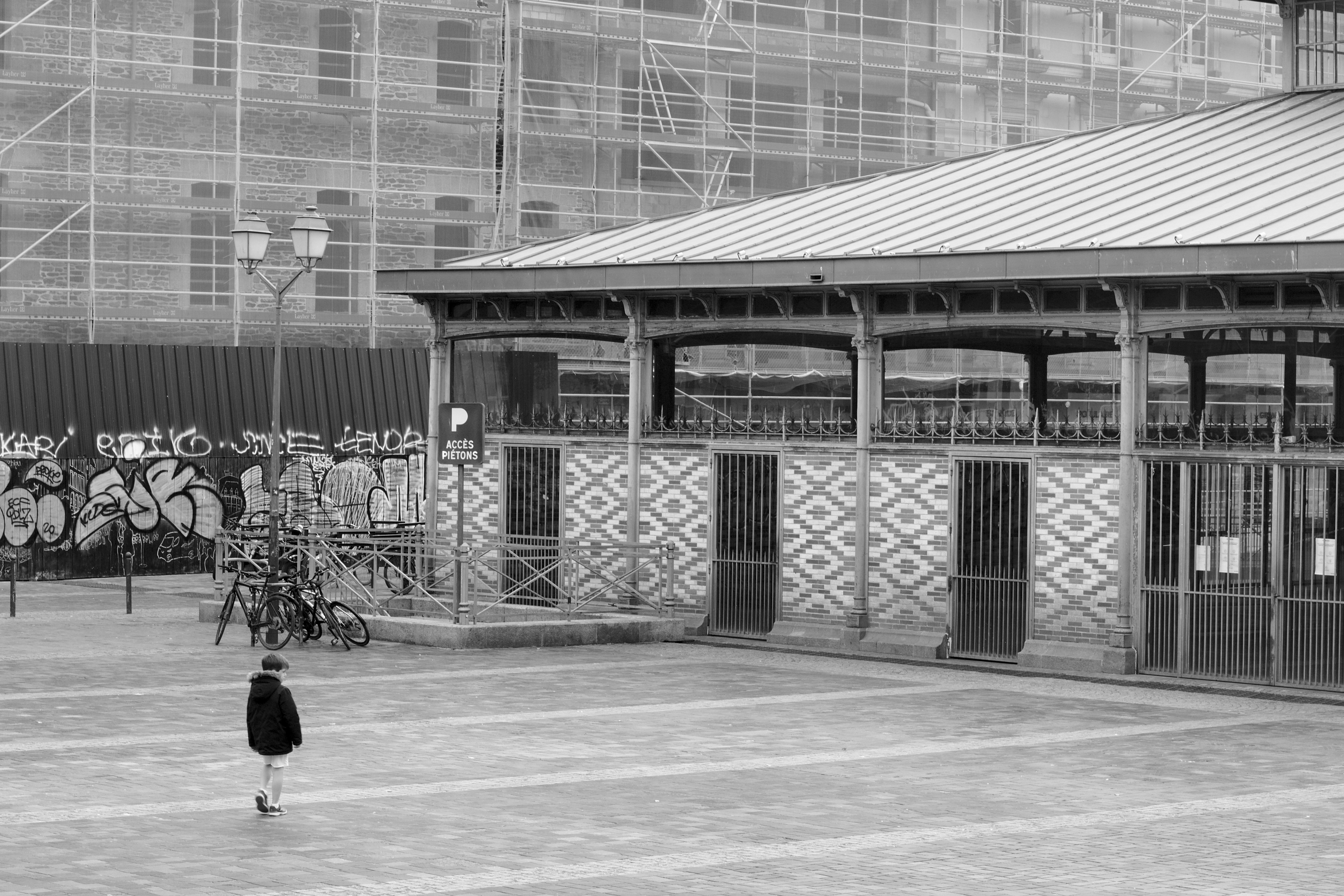 Photo en noir et blanc d'une place pavée où un enfant marche en regardant le bâtiment des halles qui borde la place. Contre ce bâtiment, il y a un escalier vers un parking indiqué par un panneau, et un lampadaire ancien contre lequel sont accrochés trois vélos. En fond, une palissade en métal couverte de graphes masque partiellement la façade d'un grand bâtiment en rénovation.
L'image contient beaucoup de lignes parallèles : celles en biais des pavés au sol et du toit des halles d'une part, celles verticales des colonnes sur les murs des halles, des panneaux et de l'échafaudage d'autre part.