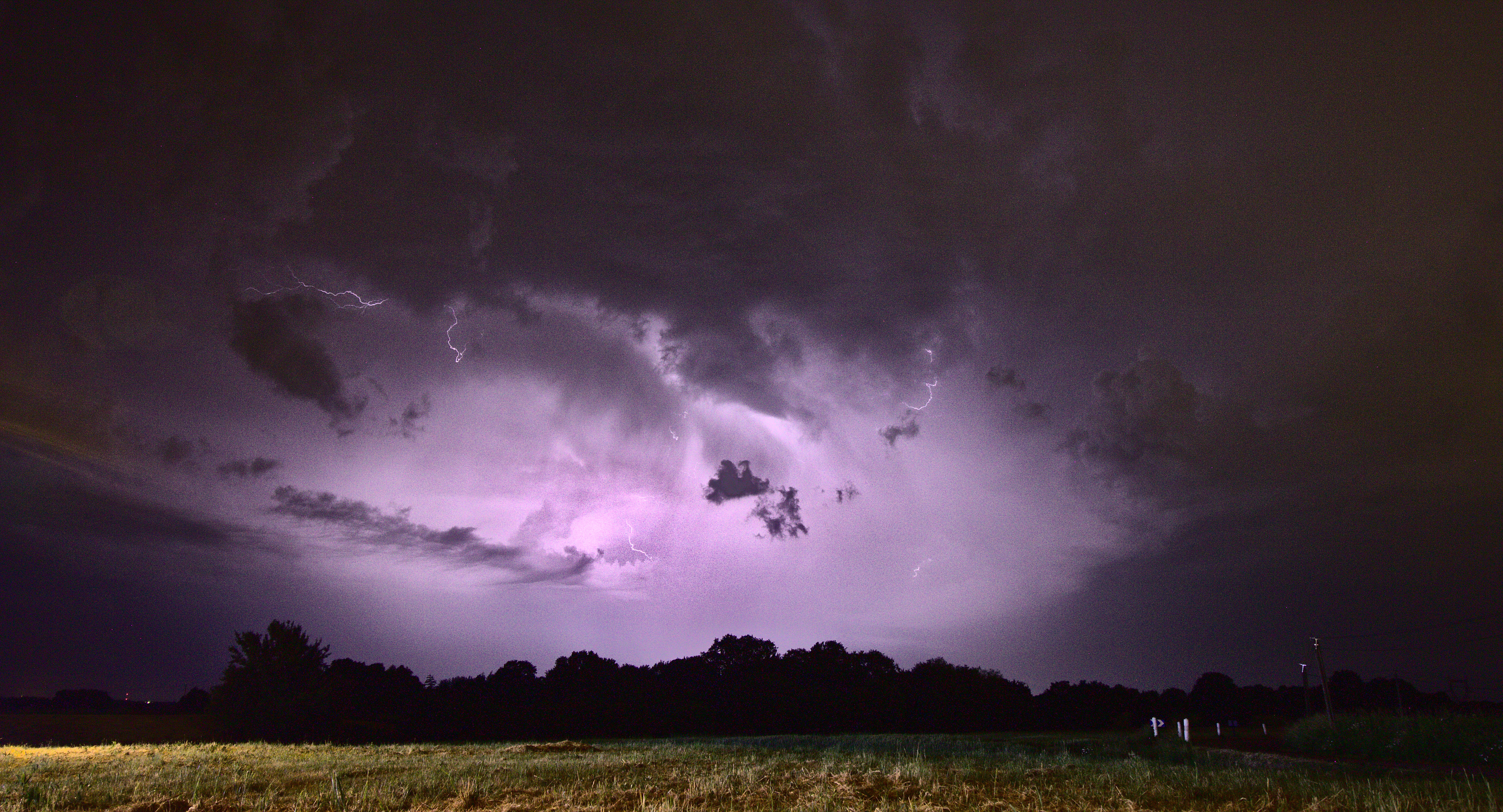 Photo de nuit montrant un terrain vague éclairé et bordé d'une haie d'arbres, au-dessus desquels un orage se déchaine. Au centre de l'image, les lourds nuages noirs tourbillonnants  sont colorés en violet par une série d'éclairs intra-nuageux, dont on peut apercevoir certains arcs électriques.