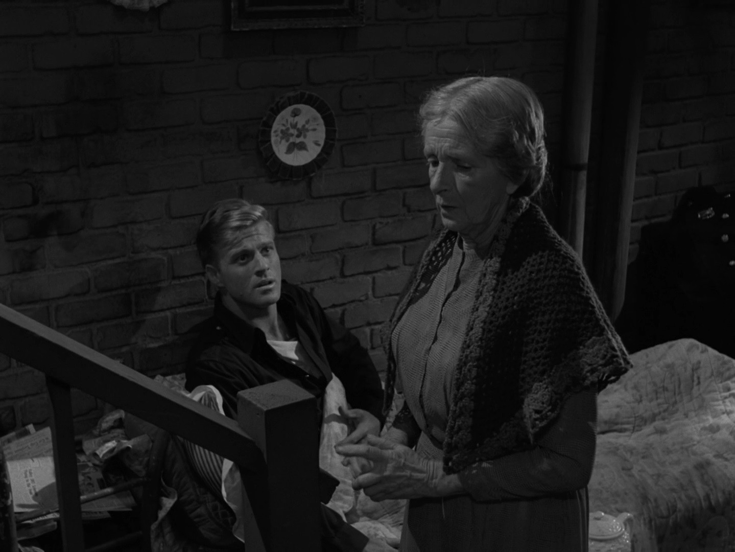 Robert Redford, assis dans un lit, sous une couverture, écoute une vieille dame debout, se tenant de profil devant lui. Elle est entrain de parler, il l'écoute attentivement.